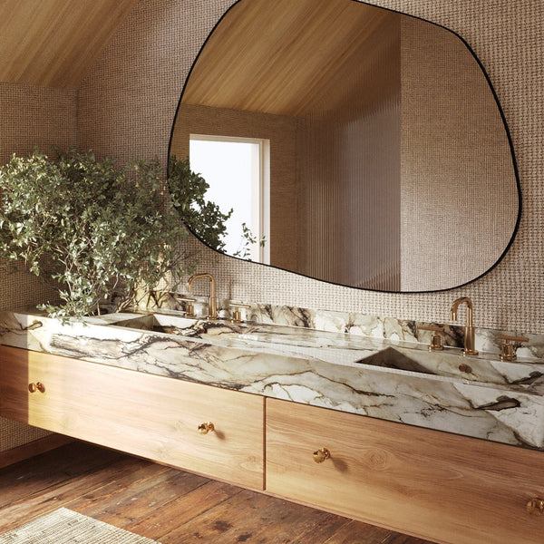 Asymmetrical wall mirror mounted above a double marble bathroom vanity with brass fixtures and wood cabinetry in a warm, naturally lit space.