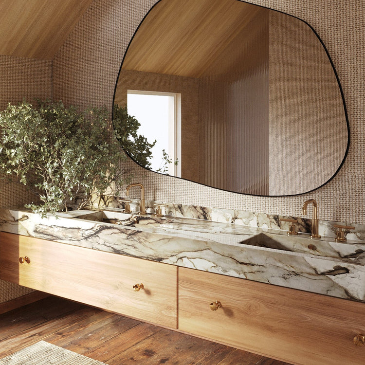 Asymmetrical wall mirror mounted above a double marble bathroom vanity with brass fixtures and wood cabinetry in a warm, naturally lit space.