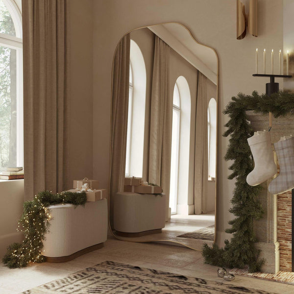 Asymmetrical full-length mirror with an organic shape and thin brass-colored frame, styled in a softly lit living space with holiday decor and arched windows.
