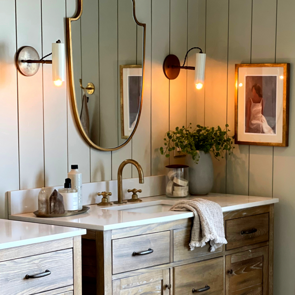 Shield-shaped wall mirror with a gold frame, mounted vertically above a bathroom vanity between two wall sconces.