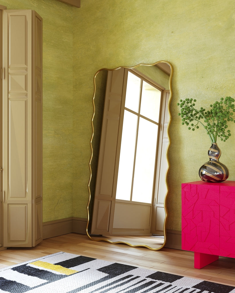 Full-length rectangular mirror with a gold frame and softly wavy edges, leaning vertically against a wall in a living space.