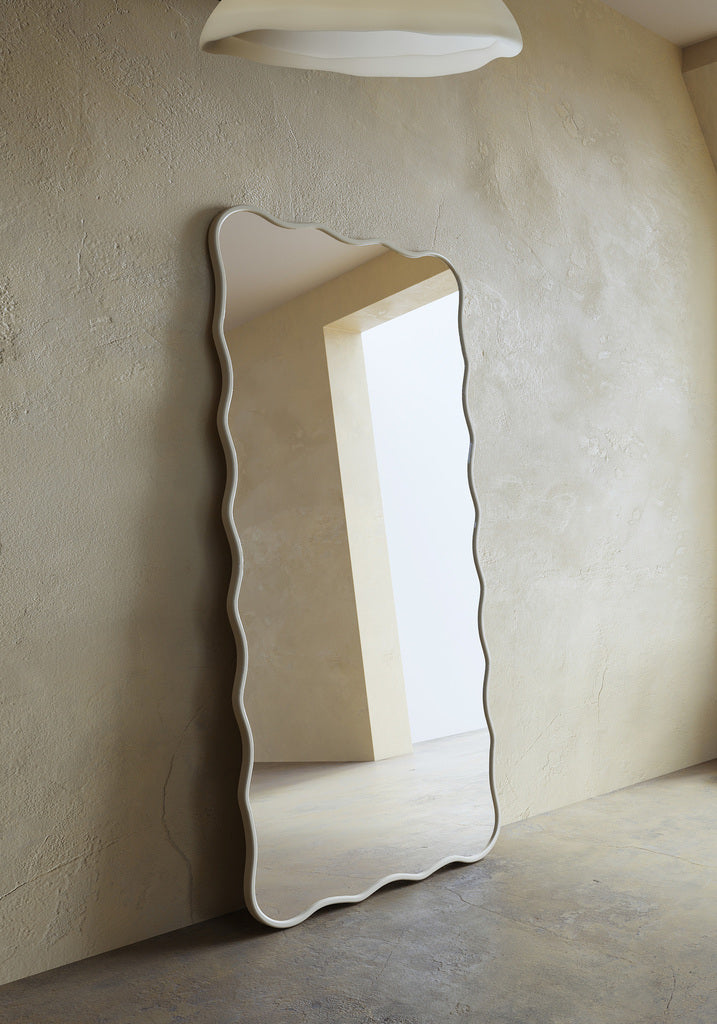 Full-length rectangular mirror with softly wavy edges and a beige cream colored frame, leaning vertically against a wall.