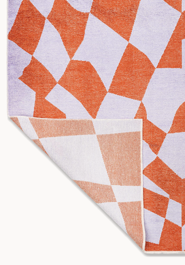 Corner detail of a flat-woven rug with rust and lavender geometric pattern.