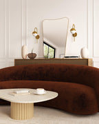 Living room interior with a curved brown sofa, round light-colored coffee table, wall mirror, and two brass wall sconces with white glass shades.