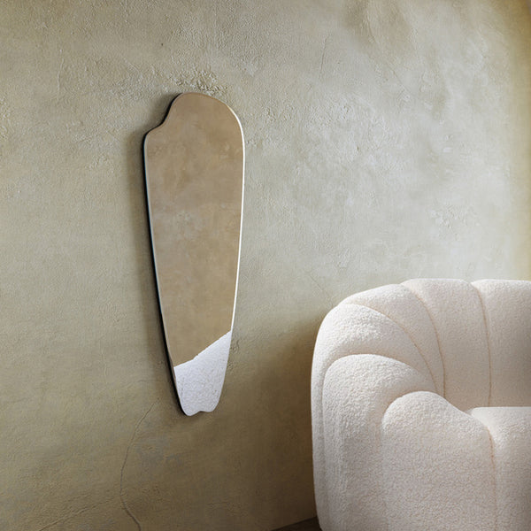 Frameless elongated mirror with an organic silhouette mounted on a textured wall beside a curved upholstered chair.