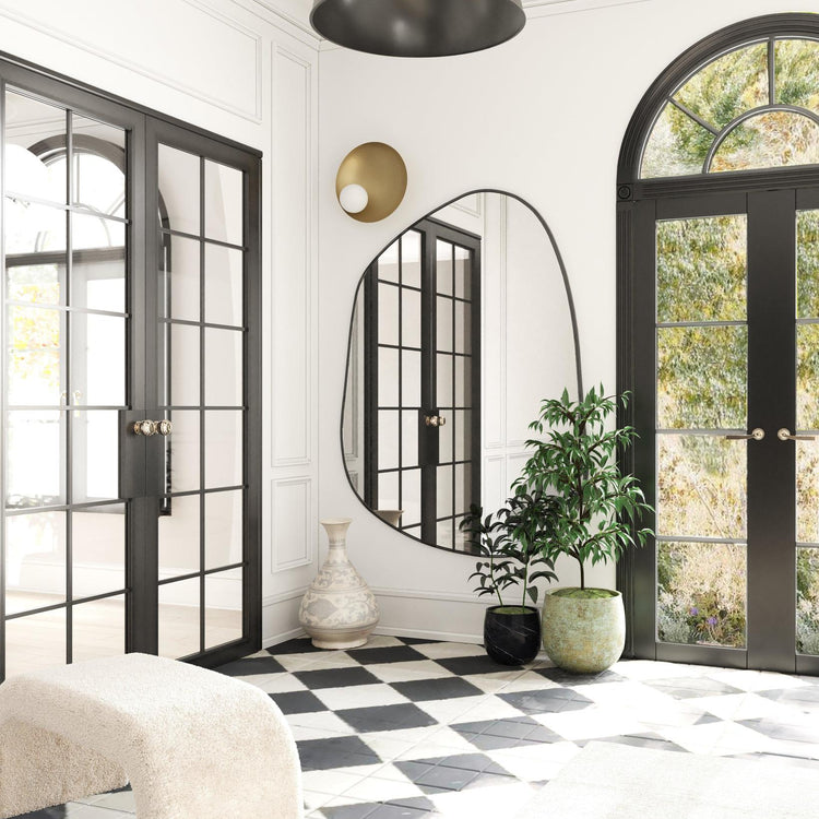 Asymmetrical wall mirror mounted in a bright entryway with black-framed doors, patterned tile flooring, and potted plants.