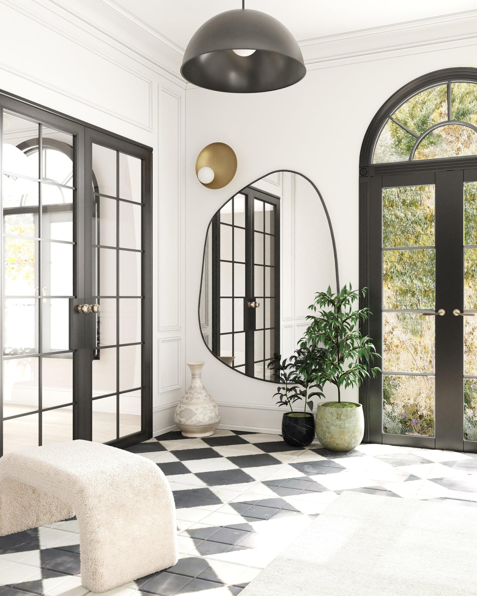 Asymmetrical wall mirror mounted in a bright entryway with black-framed doors, patterned tile flooring, and potted plants.