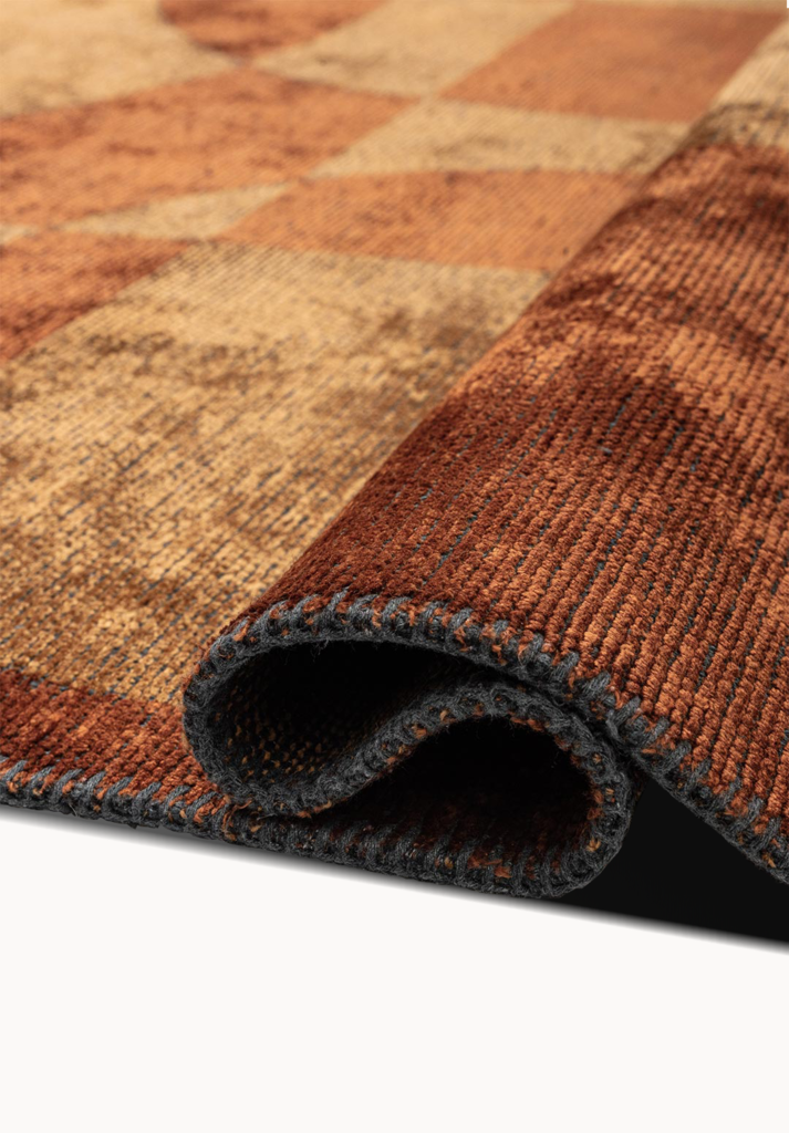 Close-up of a flat-woven rug corner folded over, highlighting the stitched edge and textured weave in warm caramel, rust, and tan tones with a subtly distressed, earthy finish.