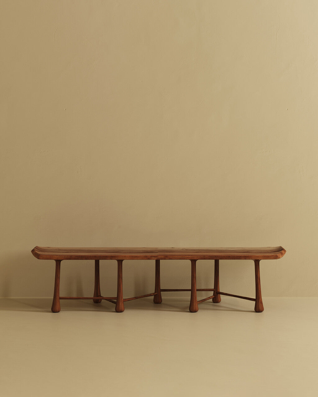 A long wooden bench with a gently curved seat and multiple rounded legs, positioned against a plain wall on a neutral floor.