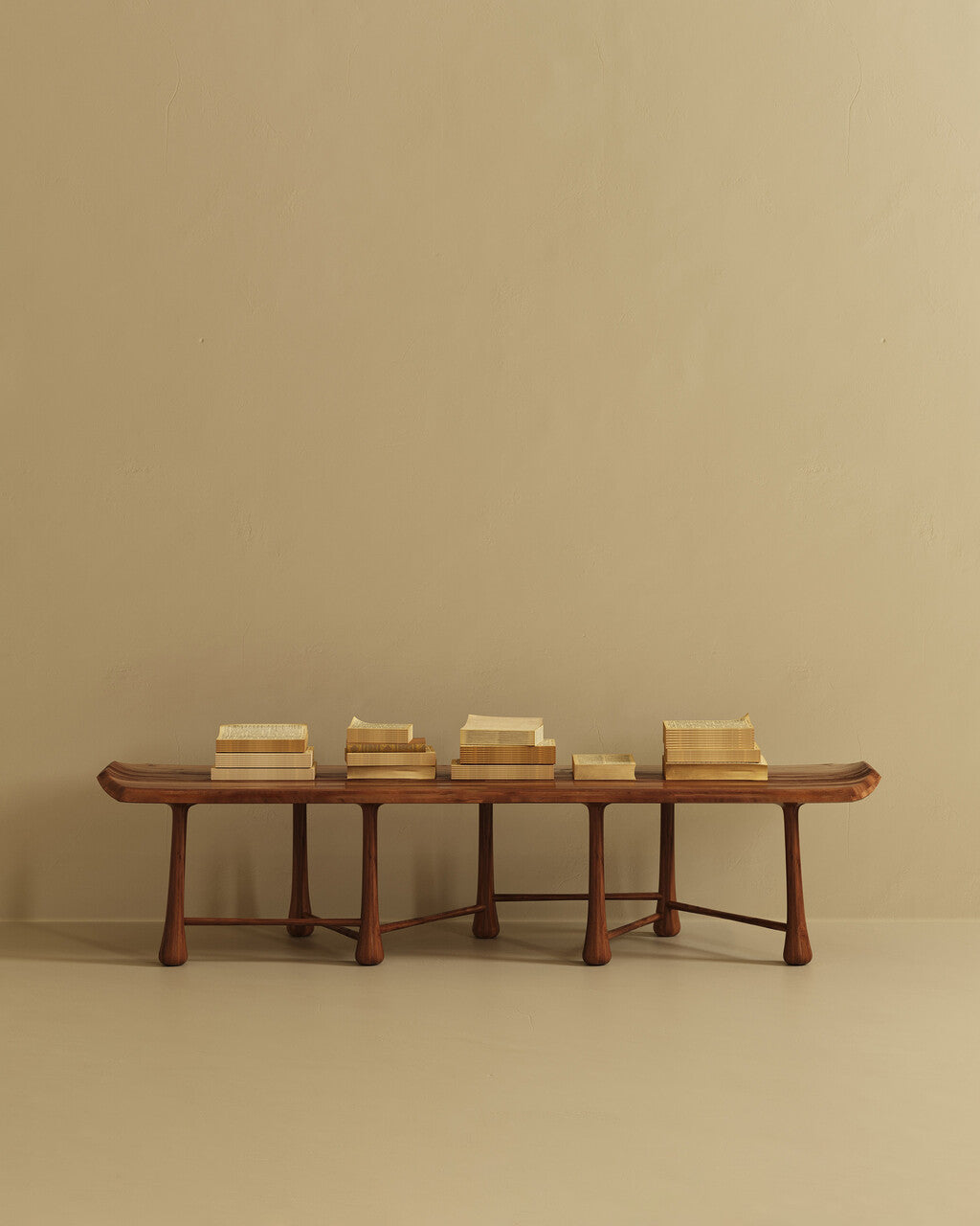 A long wooden bench with a gently contoured seat and multiple rounded legs, placed against a plain wall, with several stacked books arranged along the top.