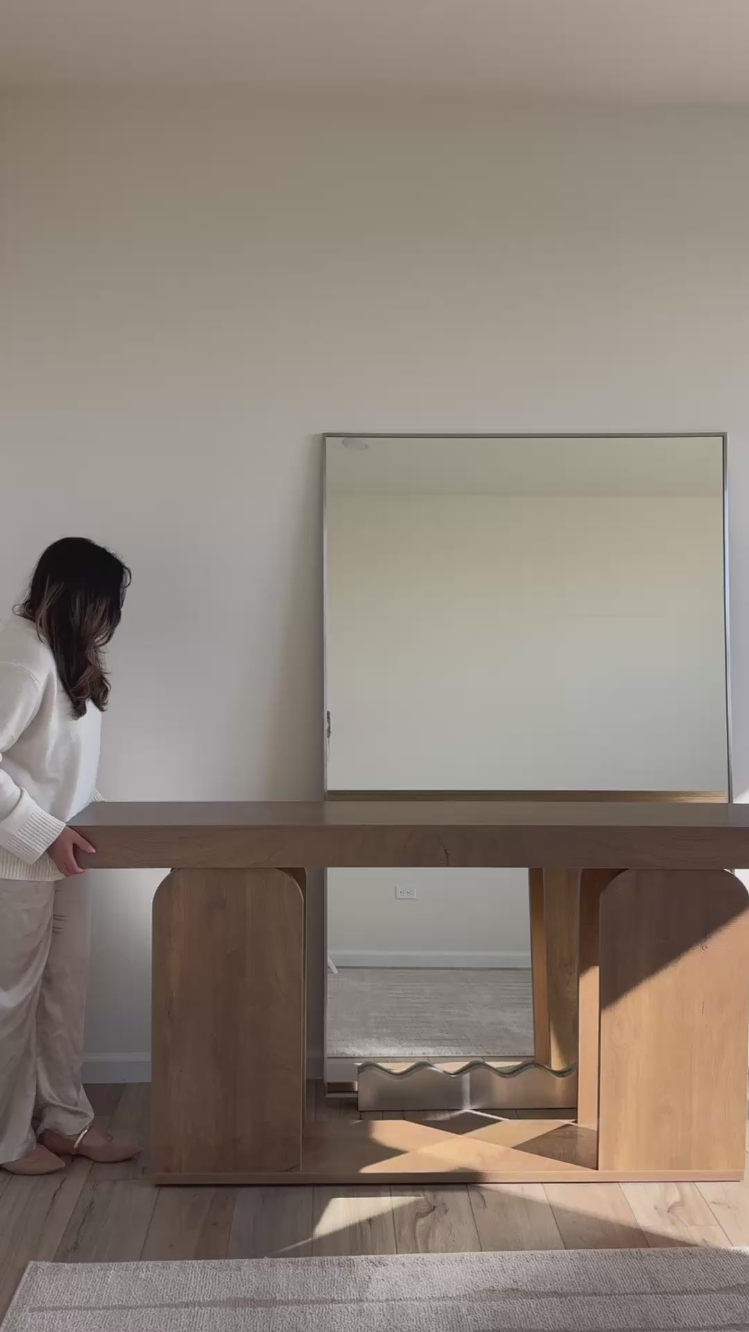 A woman decorates a full-length rectangular mirror placed behind a console table, styling the space with holiday season accents in a warm, softly lit interior.