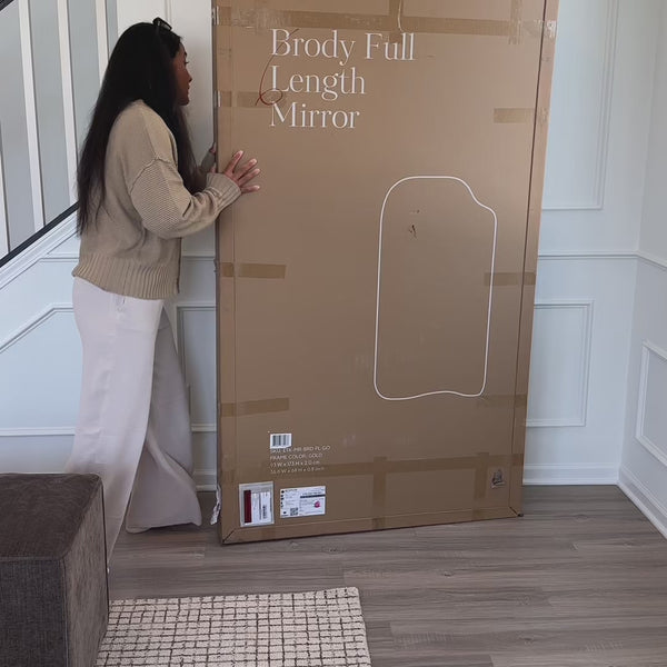 A woman unboxes a full-length mirror and places it in a living room, with natural daylight illuminating the space and reflecting in the mirror as it is positioned.