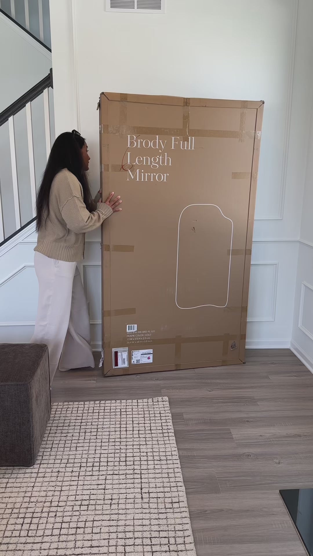 A woman unboxes a full-length mirror and places it in a living room, with natural daylight illuminating the space and reflecting in the mirror as it is positioned.